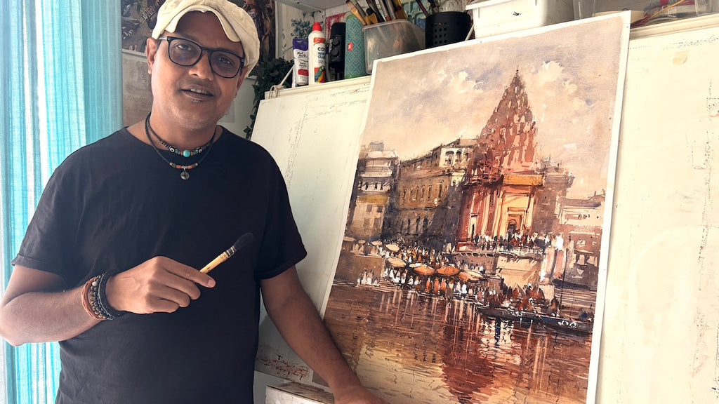 watercolor artist nitin singh holding a paintbrush next to a large painting of a varanasi ghat scene